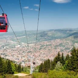 TRAVELSAGE SPÓŁKA Z OGRANICZONĄ ODPOWIEDZIALNOŚCIĄ - Czerwona kabina kolejki linowej z pasażerami na tle panoramy miasta i gór, widok zalesionego zbocza. Błękitne niebo, słoneczny dzień.