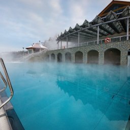 TRAVELSAGE SPÓŁKA Z OGRANICZONĄ ODPOWIEDZIALNOŚCIĄ - Zewnętrzny basen termalny z parującą wodą w zimowej scenerii. Widoczne schody ze stali nierdzewnej, budynek z kamiennymi łukami i ratunkowe koło.