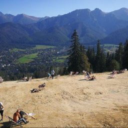 TRAVELSAGE SPÓŁKA Z OGRANICZONĄ ODPOWIEDZIALNOŚCIĄ - Widok na Zakopane z Gubałówki. Ludzie relaksują się na leżakach, w tle panorama Tatr. Słoneczny dzień, wypoczynek na świeżym powietrzu.