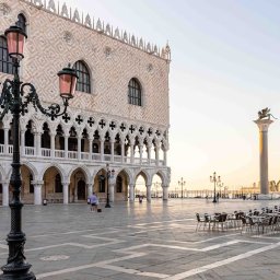 TRAVELSAGE SPÓŁKA Z OGRANICZONĄ ODPOWIEDZIALNOŚCIĄ - Plac św. Marka w Wenecji o świcie. Widok na Pałac Dożów, kolumnę ze skrzydlatym lwem i kawiarniane stoliki. Charakterystyczna latarnia na pierwszym planie.