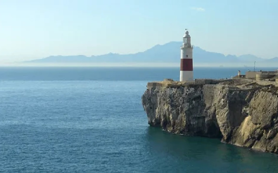 Malowniczy widok na latarnię morską Europa Point w Gibraltarze, wznoszącą się na klifie nad błękitnym morzem, z zarysem gór w oddali pod jasnym niebem.