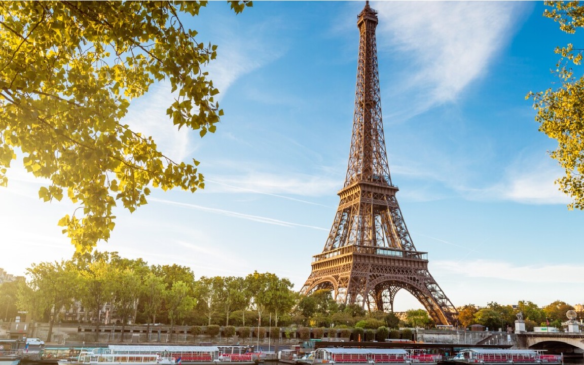 Wieża Eiffla w Paryżu, widziana z poziomu rzeki z łodziami turystycznymi. Słoneczny dzień, błękitne niebo z delikatnymi smugami chmur. Liście drzew w lewym górnym rogu.