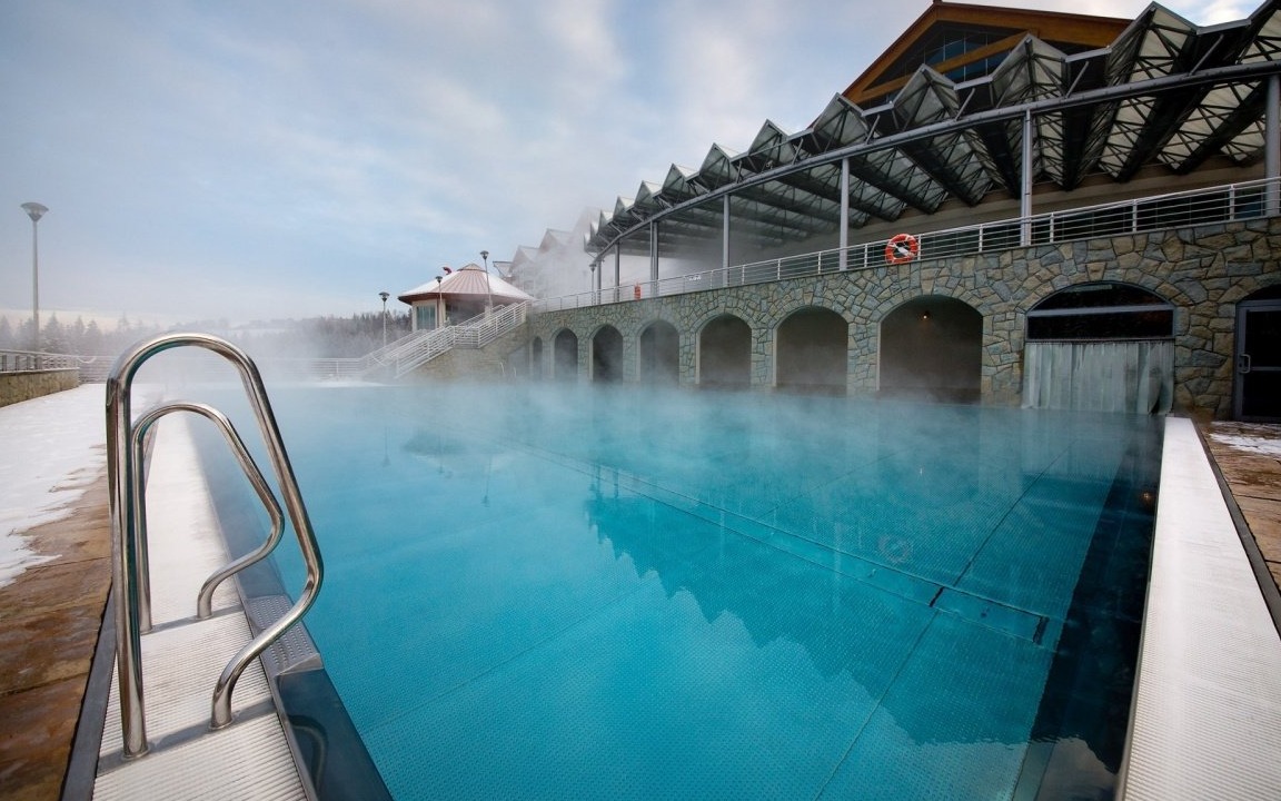 Zewnętrzny basen termalny z parującą wodą w zimowej scenerii. Widoczne schody ze stali nierdzewnej, budynek z kamiennymi łukami i ratunkowe koło.