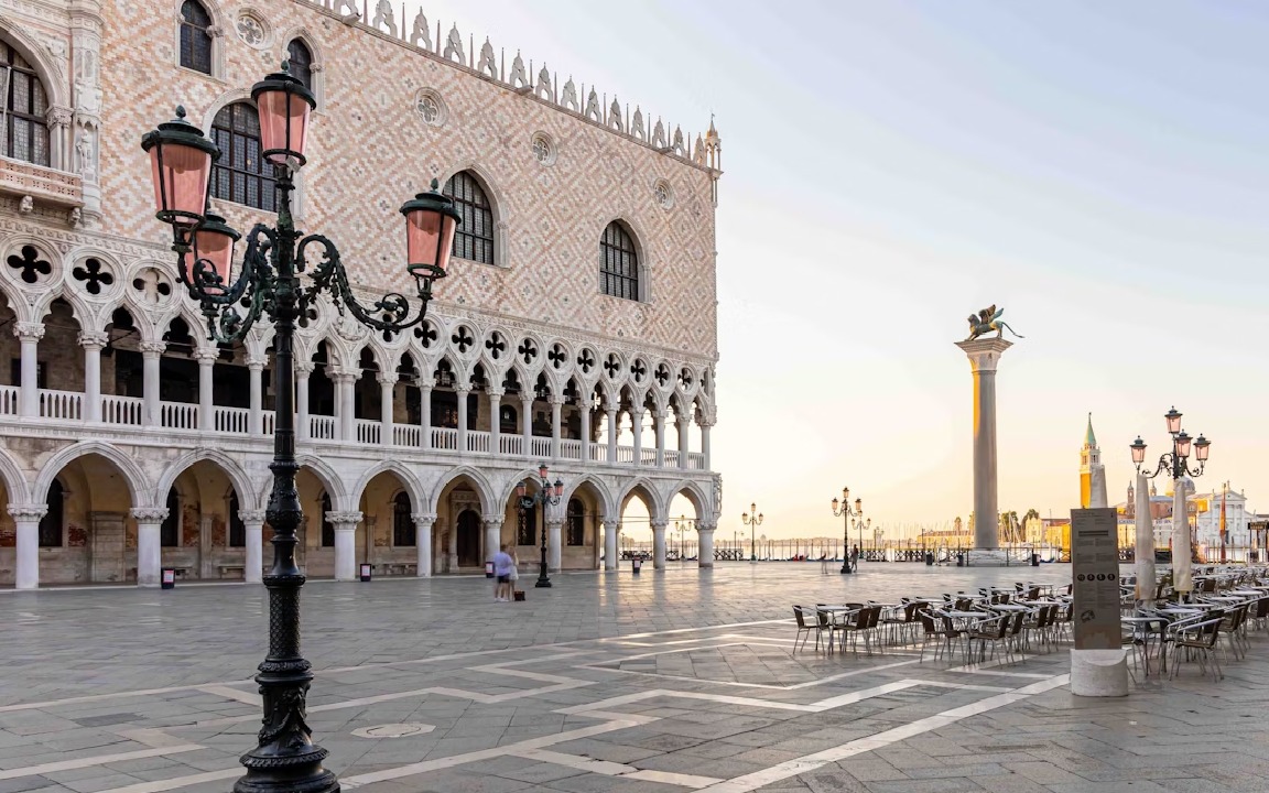 Plac św. Marka w Wenecji o świcie. Widok na Pałac Dożów, kolumnę ze skrzydlatym lwem i kawiarniane stoliki. Charakterystyczna latarnia na pierwszym planie.
