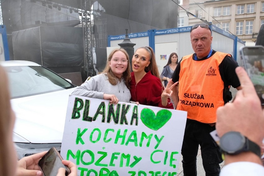 Mężczyzna w pomarańczowej kamizelce Służby Porządkowej pilnuje bezpieczeństwa podczas spotkania fanów z piosenkarką Blanką, która pozuje do zdjęcia z fanką trzymającą transparent z napisem 'Blanka...