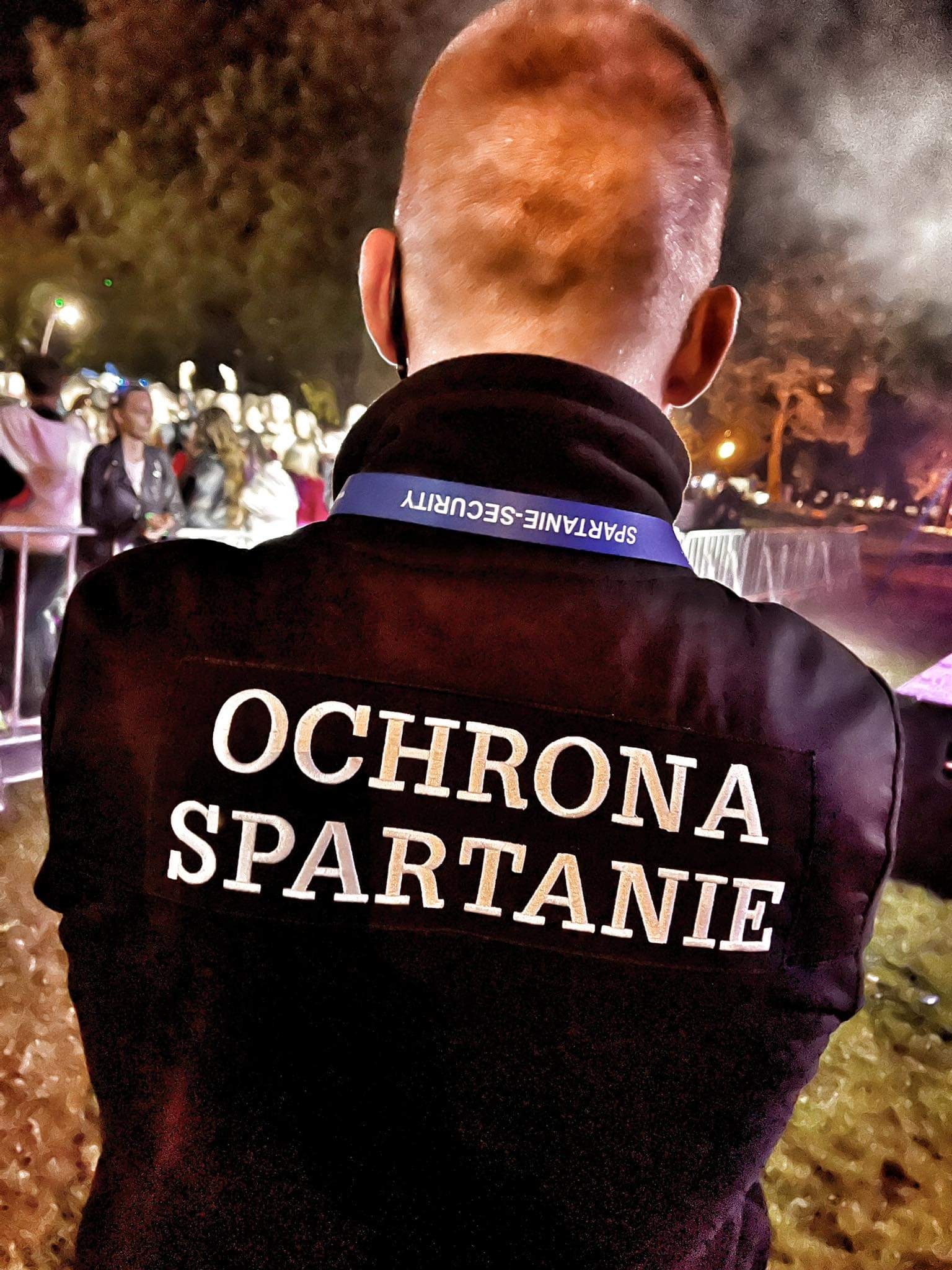 Ochroniarz tyłem, z napisem 'OCHRONA SPARTANIE' na kurtce, w tle widoczny tłum ludzi i oświetlenie sceniczne.
