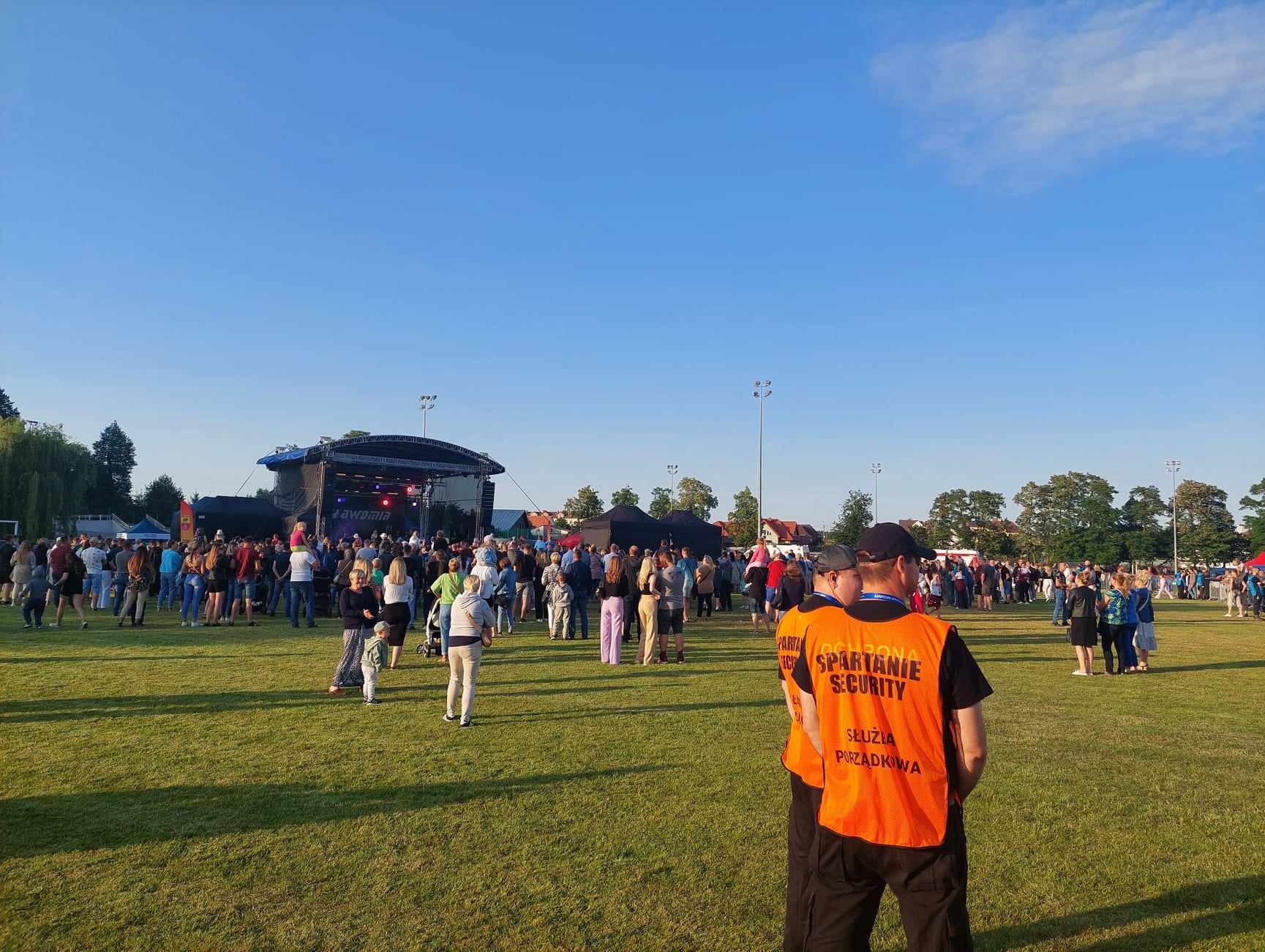 Dwóch ochroniarzy w pomarańczowych kamizelkach z napisem 'Spartanie Security' stoi tyłem do kamery, obserwując duży plenerowy koncert na trawiastym terenie.