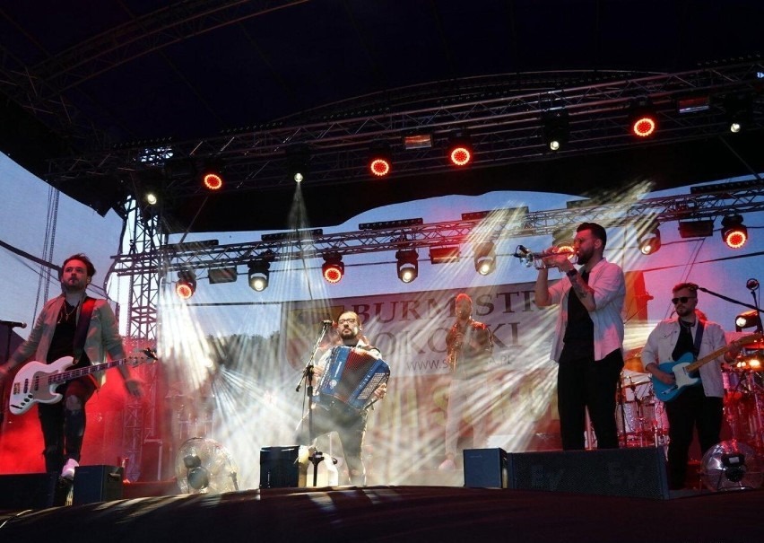 Zespół muzyczny grający na scenie podczas koncertu: gitarzysta basowy, akordeonista, trębacz i gitarzysta elektryczny w blasku reflektorów.