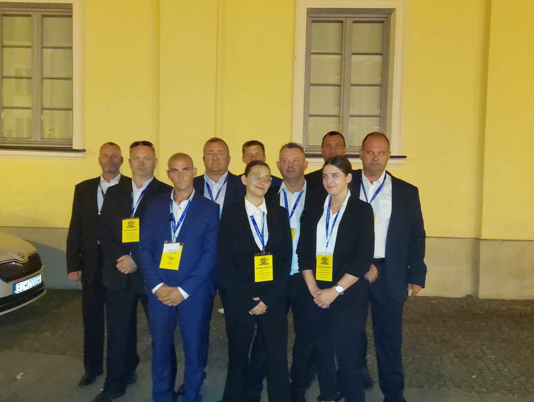 Grupa dziewięciu osób w garniturach i z identyfikatorami na szyi, stojących przed żółtą ścianą budynku z dwoma oknami, przy zaparkowanym samochodzie marki Skoda.