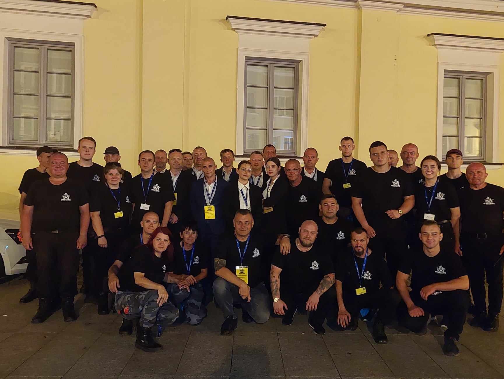 Grupowe zdjęcie pracowników firmy ochroniarskiej z Białegostoku w czarnych koszulkach z logo firmy, stojących i kucających przed budynkiem z żółtą elewacją i oknami.