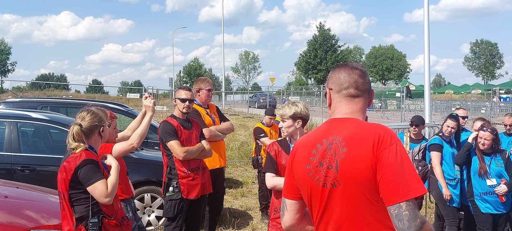 Grupa pracowników ochrony ubrana w czerwone i pomarańczowe kamizelki, stojąca na zewnątrz podczas wydarzenia plenerowego, z zaparkowanymi samochodami i drzewami w tle.