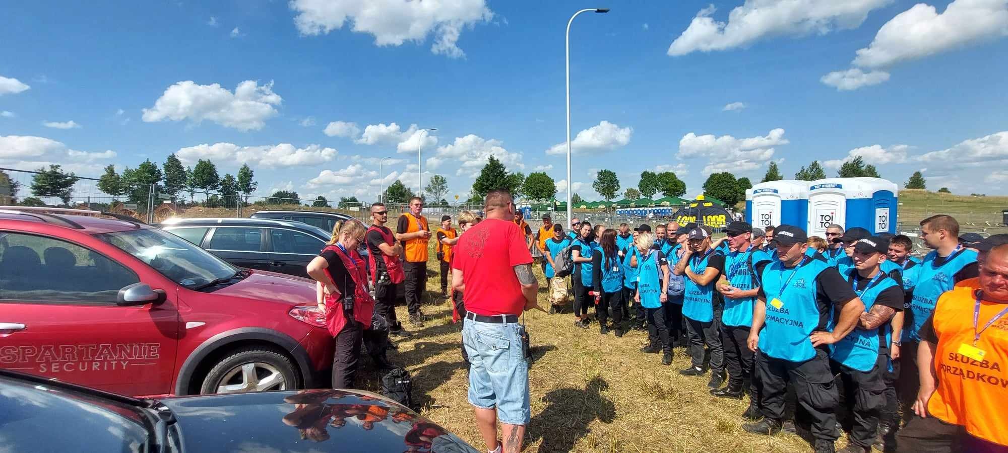 Grupa pracowników ochrony w niebieskich kamizelkach stoi na zewnątrz, słuchając instrukcji mężczyzny w czerwonej koszulce, na pierwszym planie samochód z napisem 'Spartanie Security', w tle...