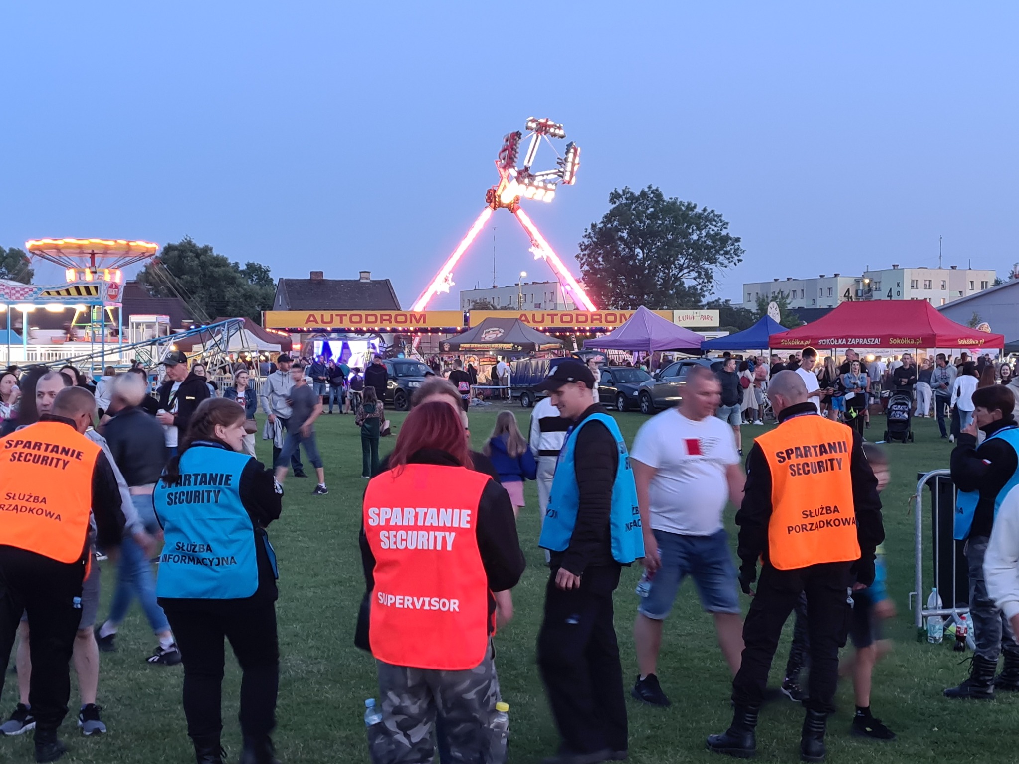 Ochrona imprezy plenerowej: ochroniarze w kamizelkach 'Spartanie Security' patrolują teren w tle wesołego miasteczka z karuzelami i autodrome.