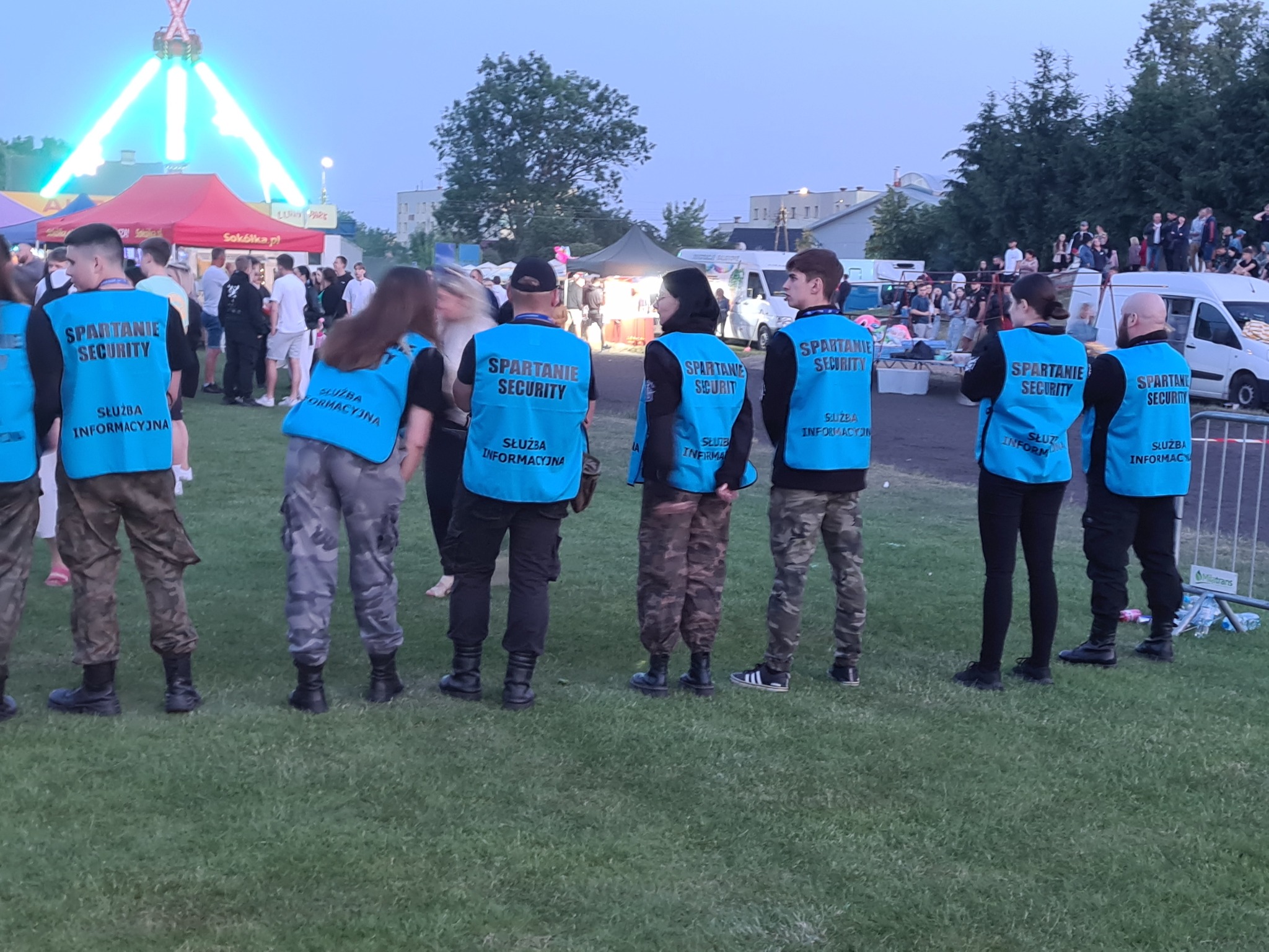 Grupa pracowników ochrony w niebieskich kamizelkach z napisem 'Spartanie Security' stoi tyłem do widza, obserwując tłum ludzi na tle wesołego miasteczka o zmierzchu.