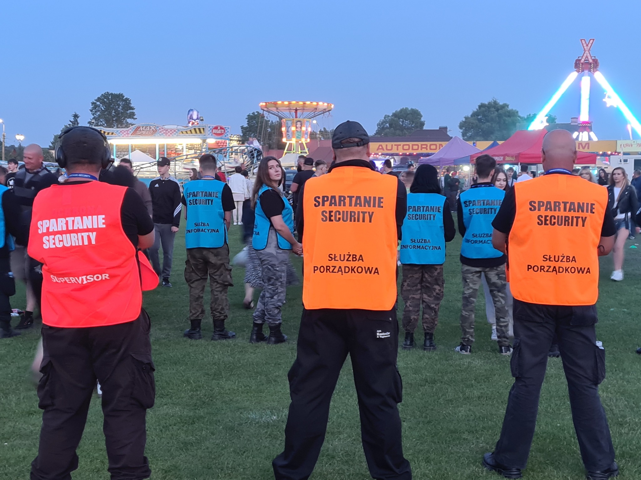 Ochrona imprezy masowej: grupa ochroniarzy firmy Spartanie Security w pomarańczowych i niebieskich kamizelkach z napisem 'Służba Porządkowa' i 'Służba Informacyjna', na tle wesołego miasteczka...