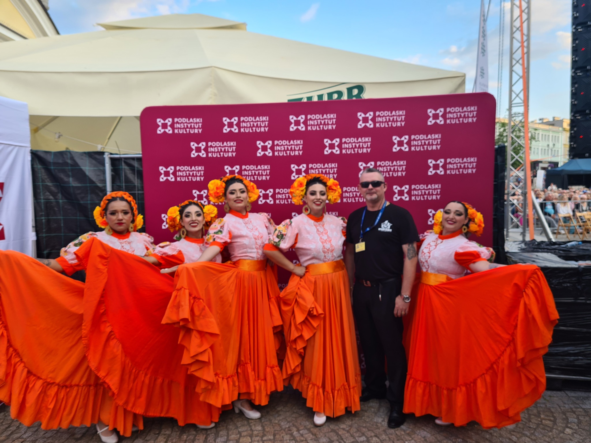 Grupa pięciu kobiet w tradycyjnych, pomarańczowych strojach tanecznych z kwiatami we włosach, pozuje na tle czerwonej ścianki z logo Podlaskiego Instytutu Kultury, obok mężczyzny w czarnym ubraniu.