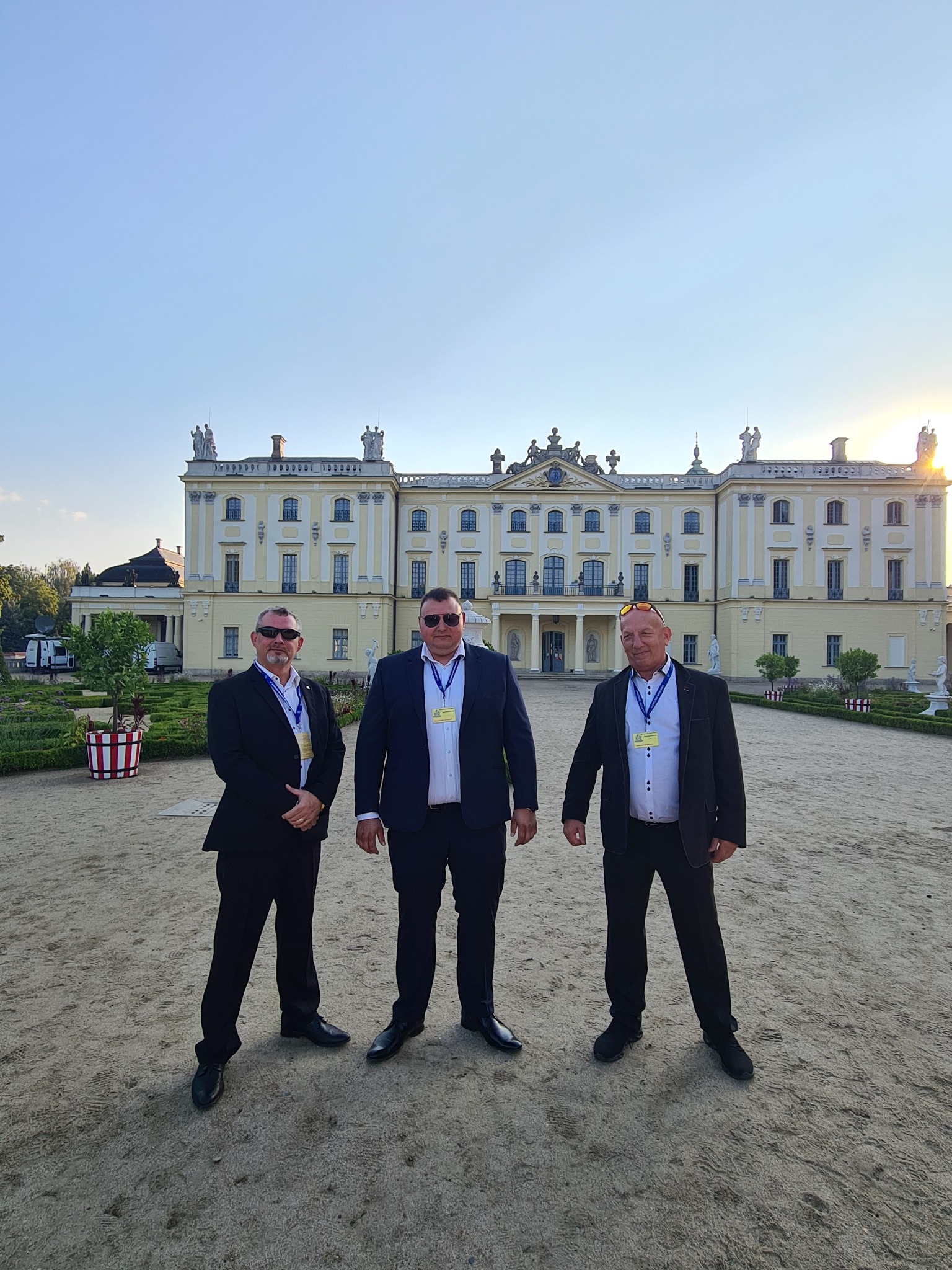 Trzech mężczyzn w garniturach z identyfikatorami na tle Pałacu Branickich w Białymstoku, jeden w okularach przeciwsłonecznych.