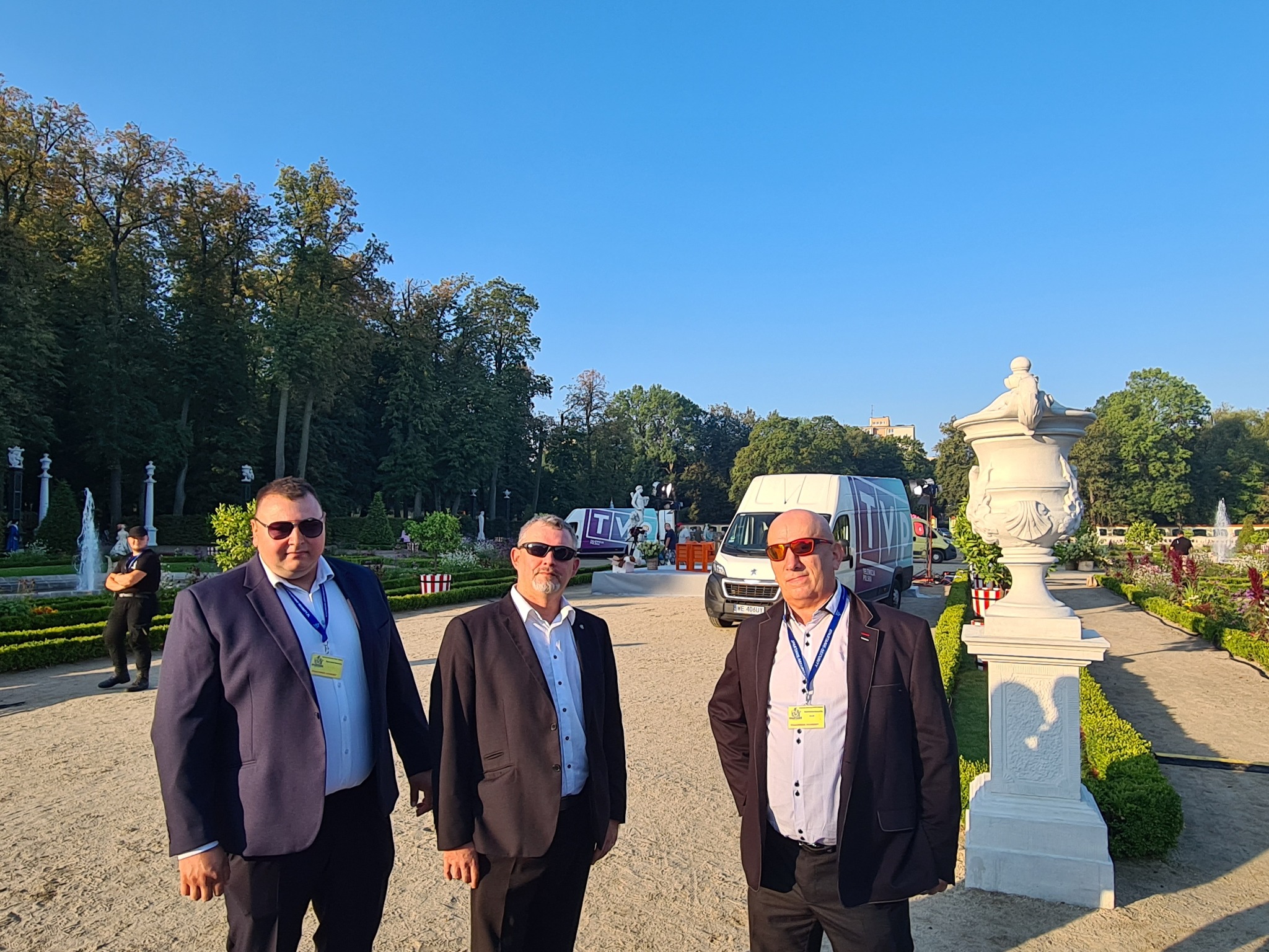 Trzech mężczyzn w garniturach i okularach przeciwsłonecznych stoi na tle parku z fontannami i zaparkowanym busem z logotypem firmy.