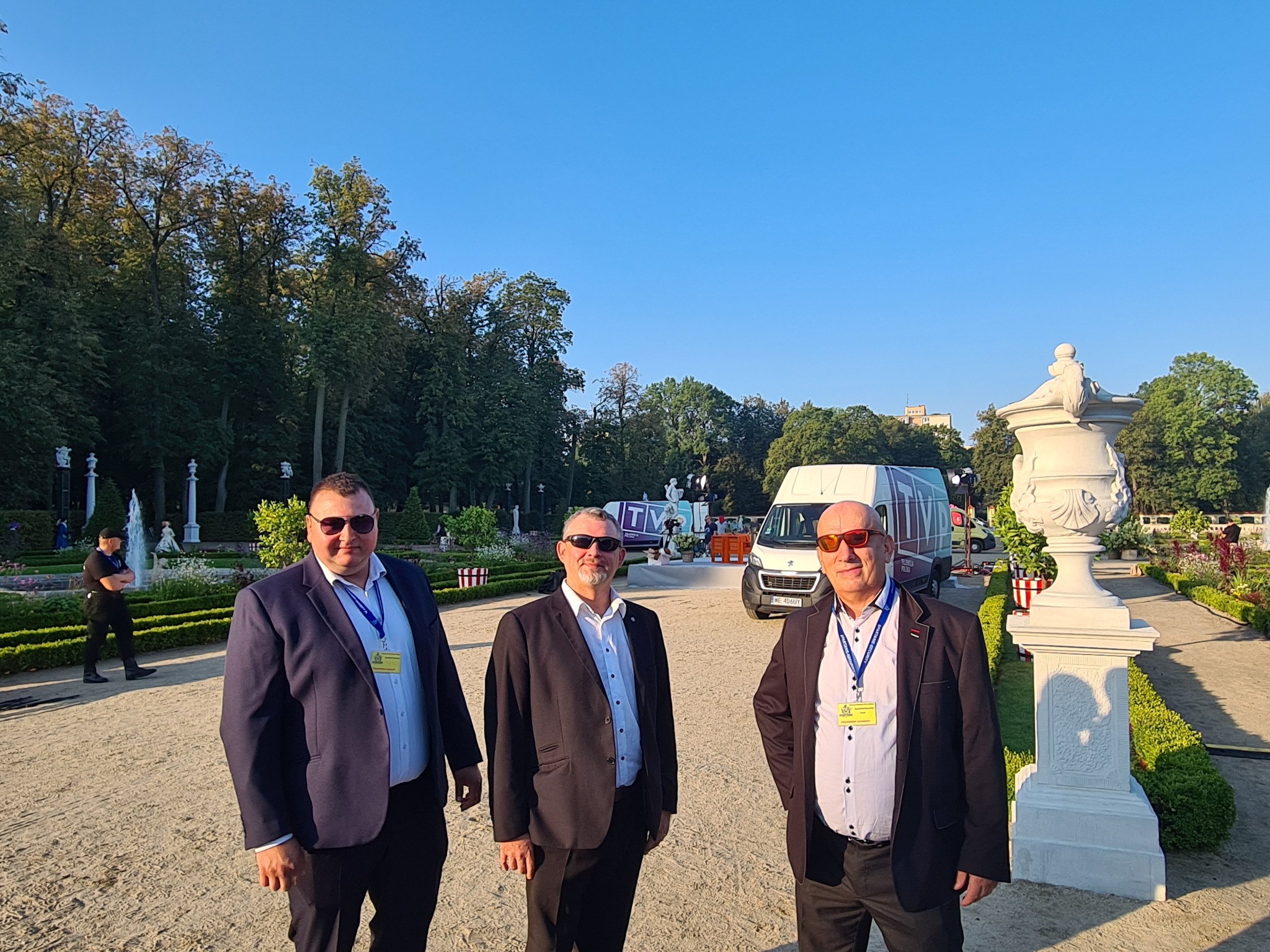 Trzech mężczyzn w garniturach i okularach przeciwsłonecznych stoi w parku, w tle fontanna i samochód dostawczy z logo TV.