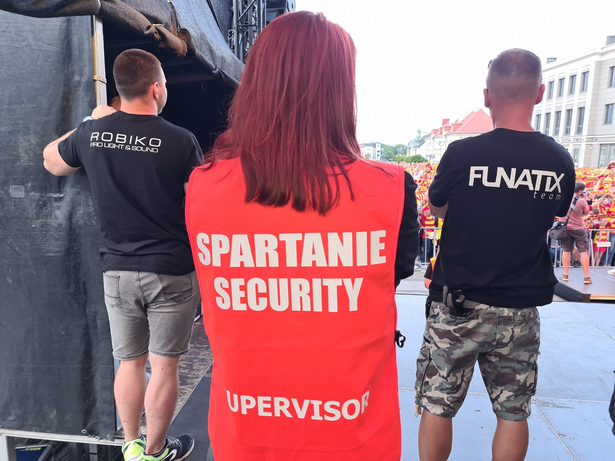 Trzech mężczyzn tyłem, w tym kobieta w czerwonej kamizelce z napisem 'Spartanie Security Supervisor', ochrona imprezy masowej w tle widoczny tłum.