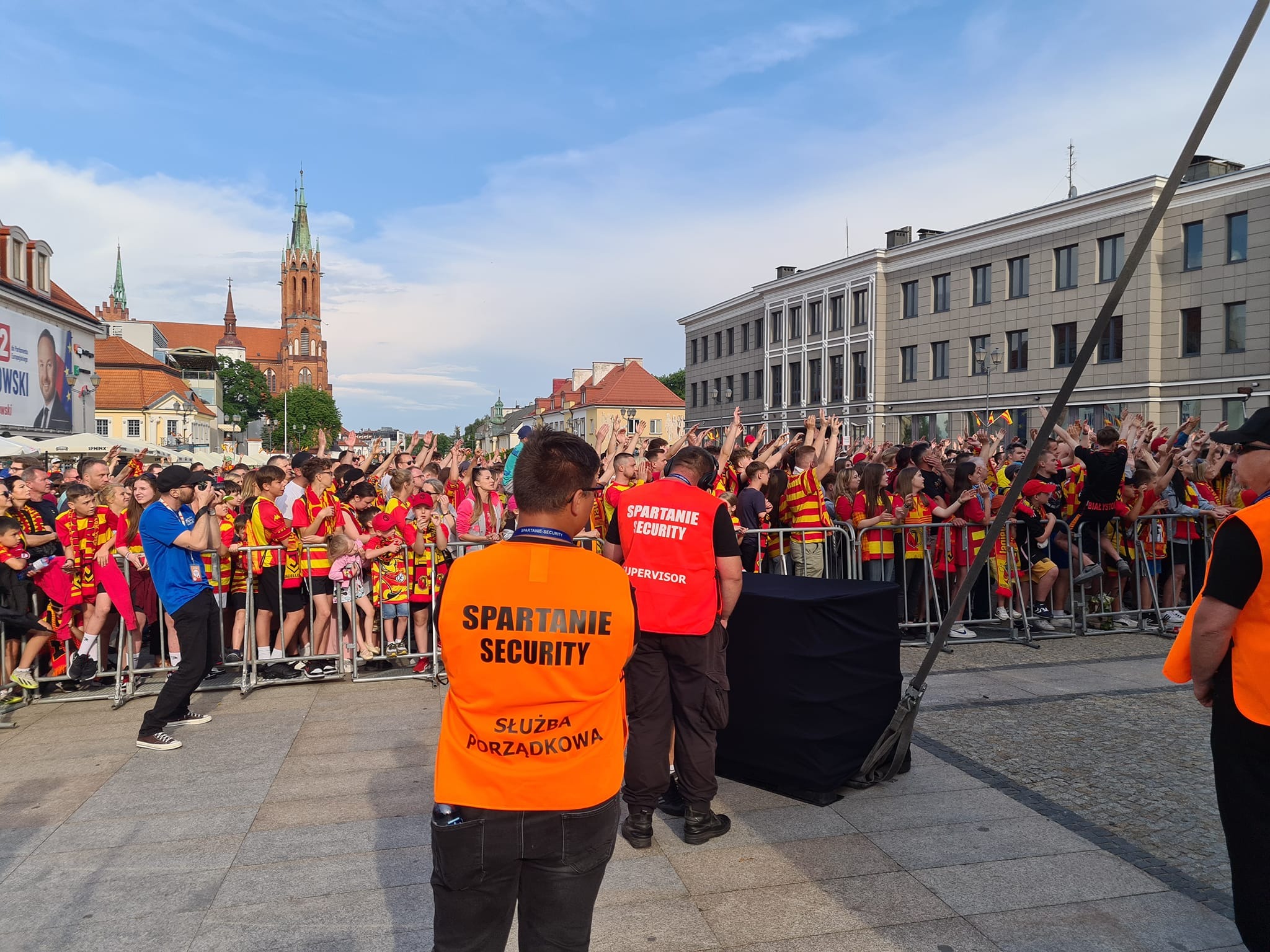 Ochrona imprezy masowej: Dwóch ochroniarzy w pomarańczowej i czerwonej kamizelce z napisem 'Spartanie Security' stoi tyłem do kamery, obserwując tłum fanów w czerwono-żółtych barwach za metalowymi...