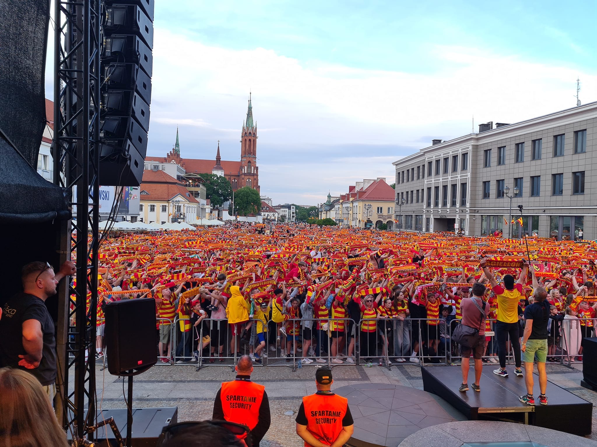 Tłum kibiców z szalikami w barwach klubowych na tle miejskiego krajobrazu, ochrona w pomarańczowych kamizelkach z napisem 'Spartanie Security' na pierwszym planie.