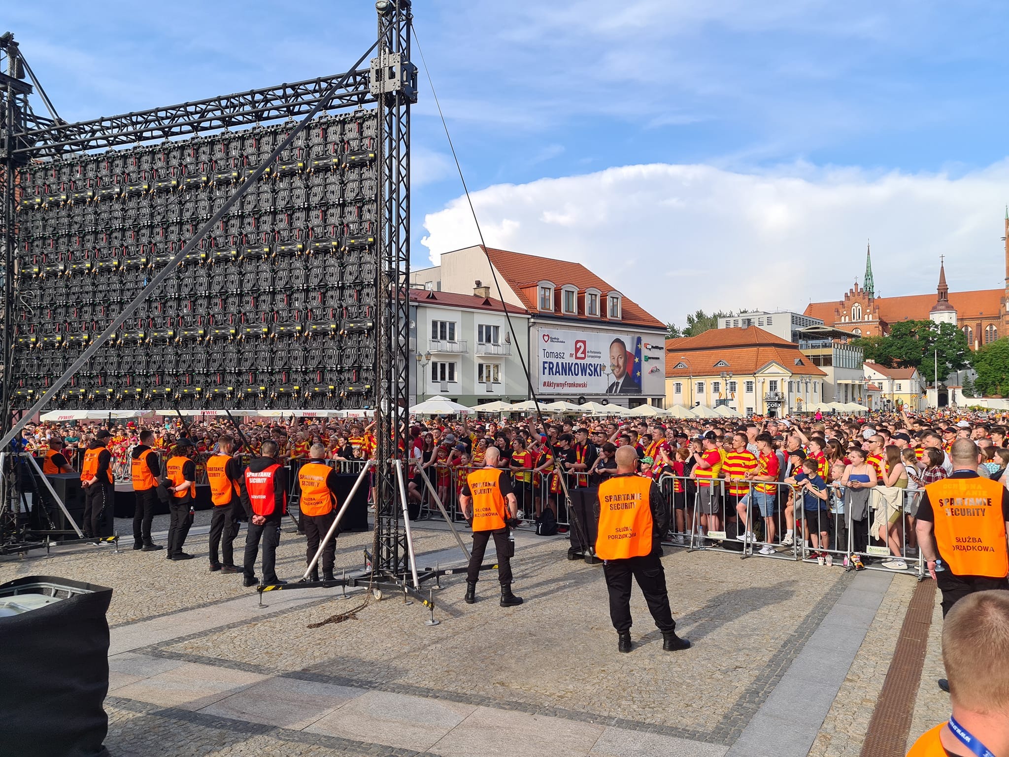 Ochrona imprezy masowej: grupa ochroniarzy w pomarańczowych kamizelkach stoi przed publicznością i telebimem podczas wydarzenia plenerowego.