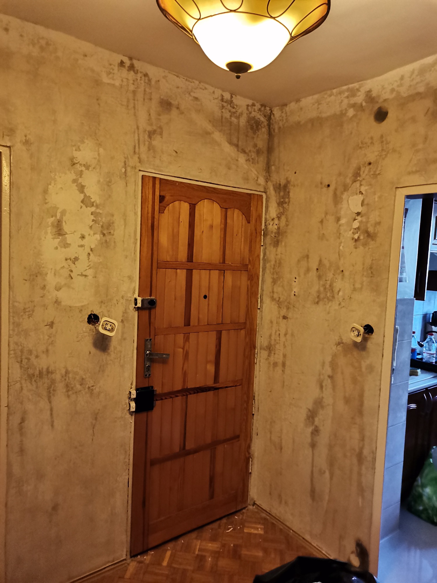 Pokój przed remontem: ściany z widocznymi śladami wilgoci i odpadającą farbą, drewniane drzwi z zamkiem, wystające gniazdka elektryczne i lampa sufitowa w stylu retro.
