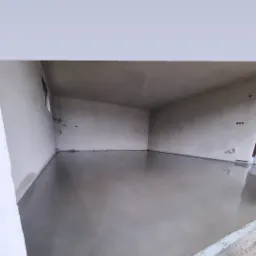 Świeżo wylana, gładka, szara posadzka w pomieszczeniu z surowymi ścianami i widocznymi instalacjami elektrycznymi.