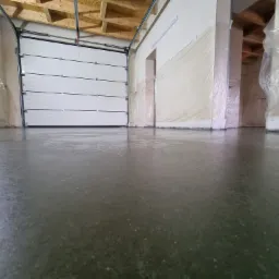 Świeżo wylana, błyszcząca, szara posadzka w hali przemysłowej z częściowo otwartą, białą bramą segmentową i zabezpieczonymi folią ścianami.