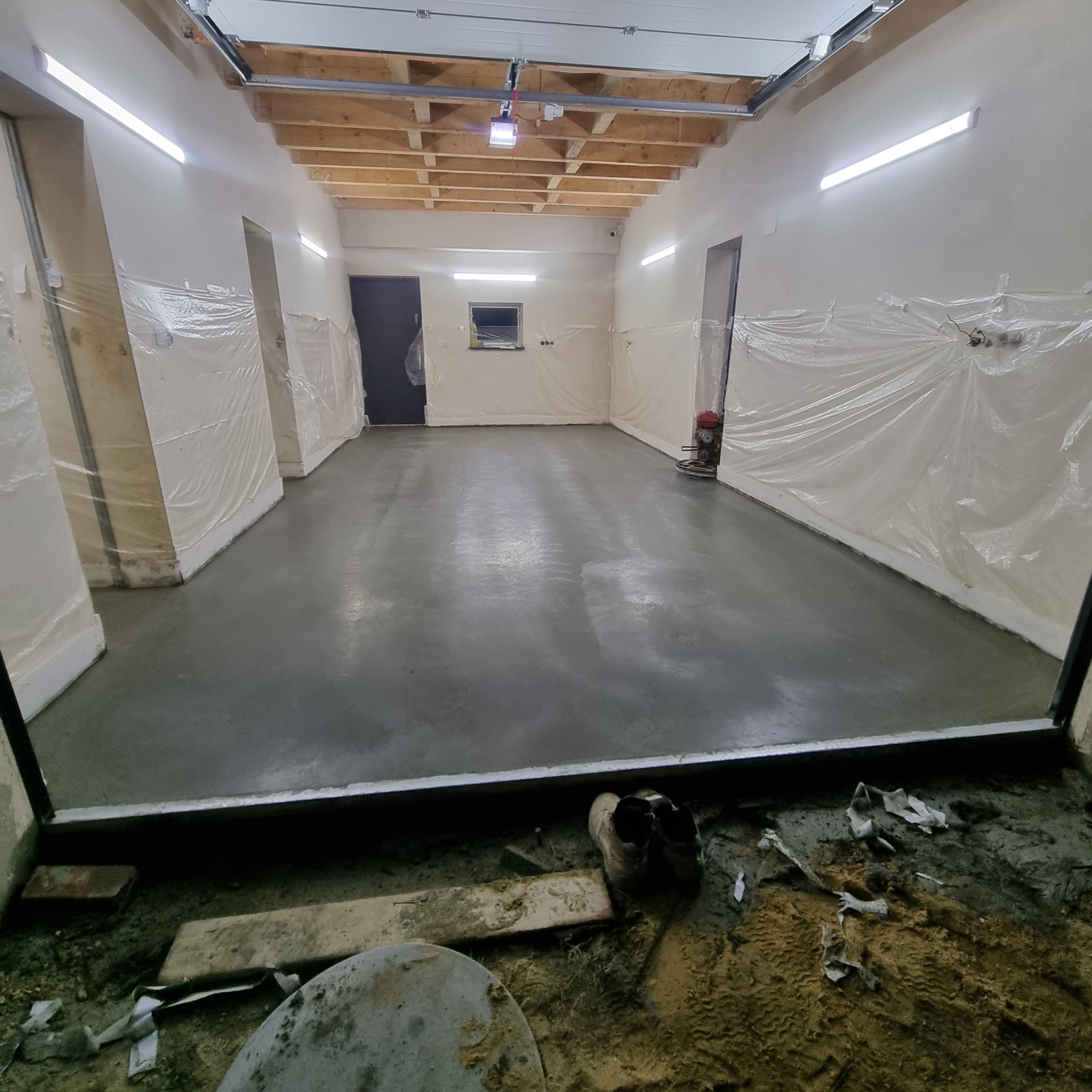 Świeżo wylana, szara posadzka w pomieszczeniu przemysłowym, ściany zabezpieczone folią, widoczna zacieraczka do betonu i robocze obuwie przy progu.