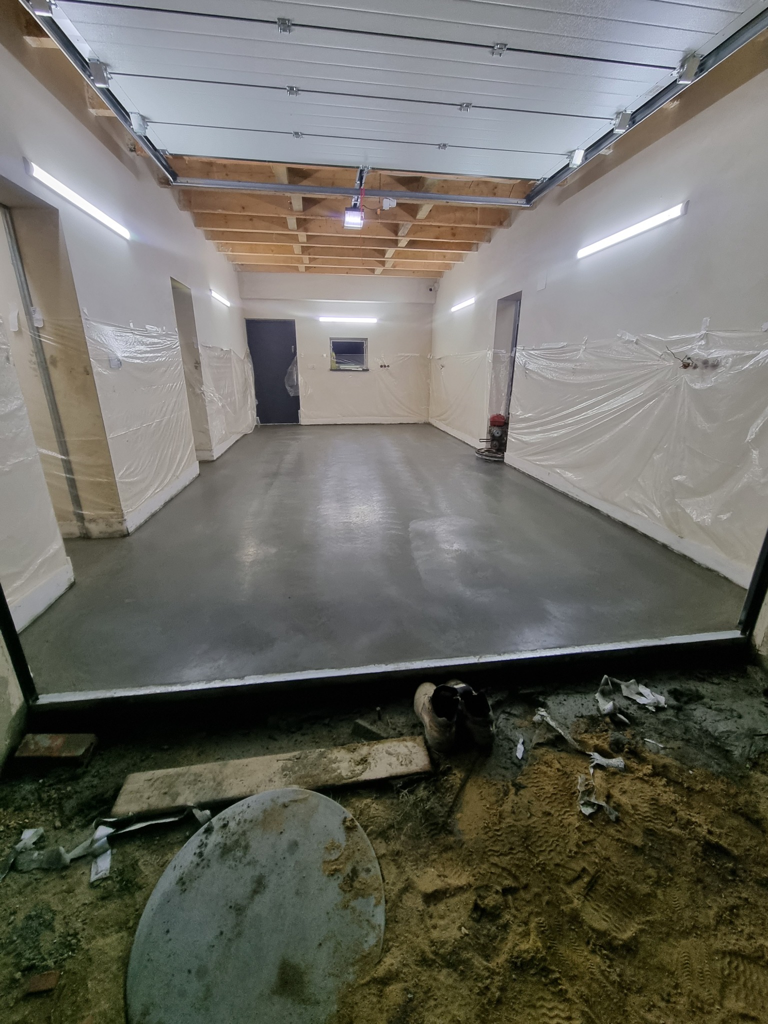 Świeżo wylana, szara posadzka w przestronnym pomieszczeniu z folią ochronną na ścianach, widoczne narzędzia i obuwie robocze w dolnym kadrze.