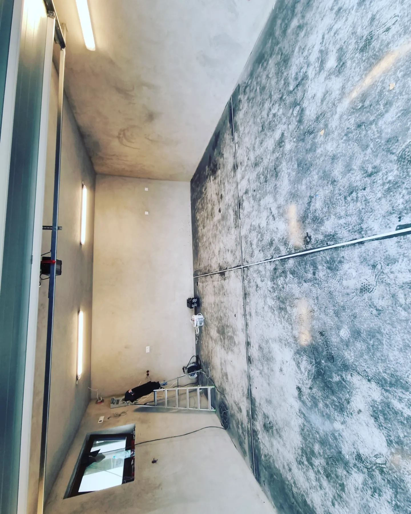 Wnętrze hali w trakcie prac wykończeniowych, z widoczną szarą, surową posadzką i ścianami o nieregularnej fakturze, drabina i sprzęt budowlany w tle, oświetlenie jarzeniowe na suficie.