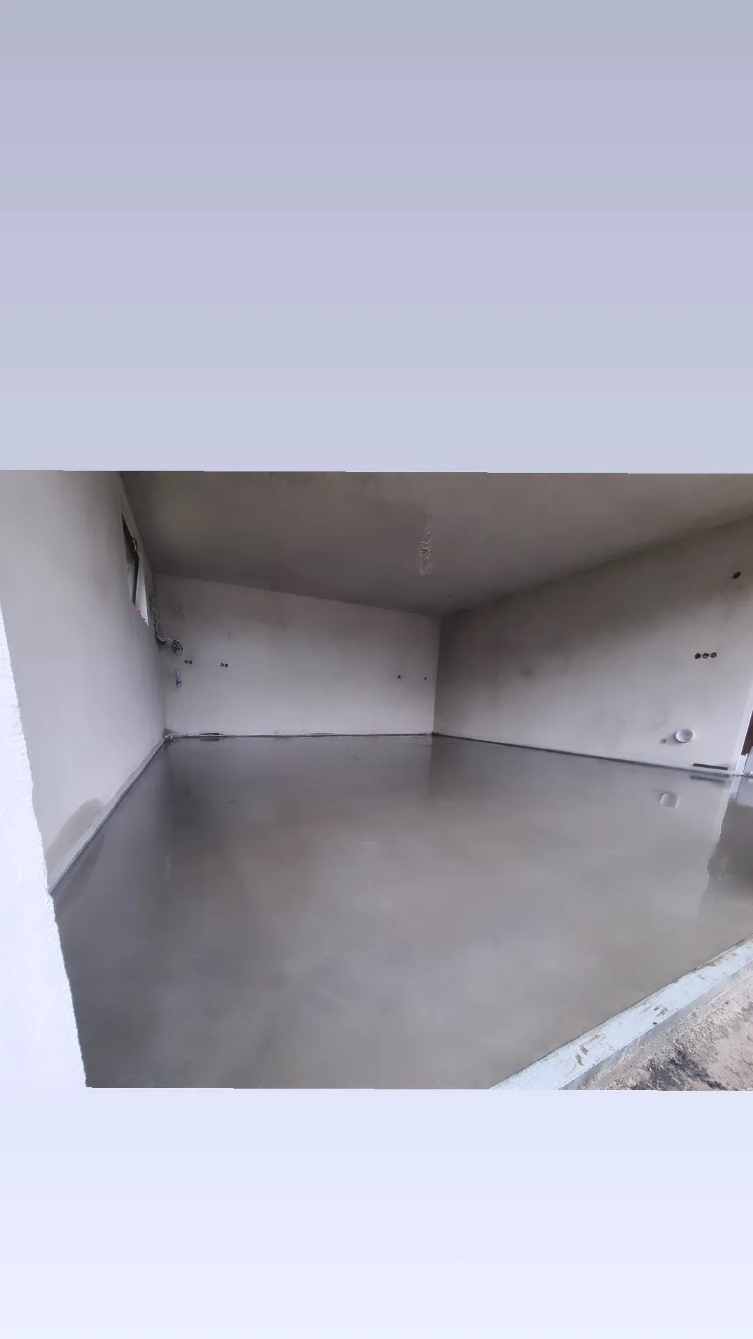 Świeżo wylana, gładka, szara posadzka w pomieszczeniu z surowymi ścianami i widocznymi instalacjami elektrycznymi.