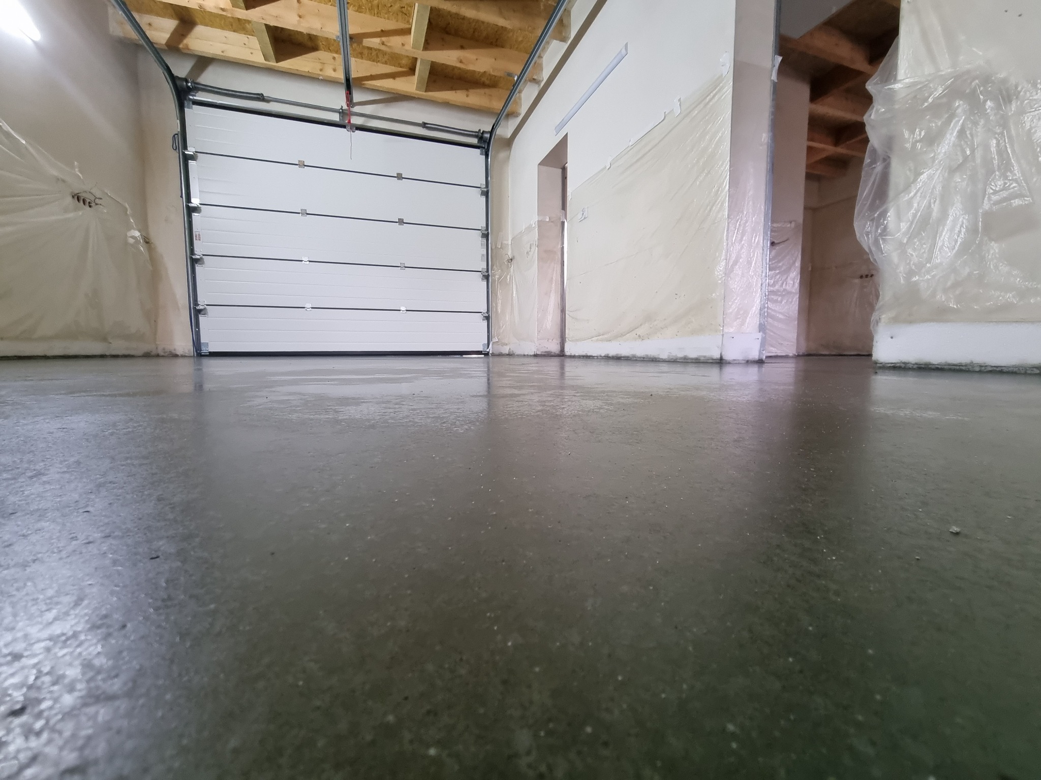 Świeżo wylana, błyszcząca, szara posadzka w hali przemysłowej z częściowo otwartą, białą bramą segmentową i zabezpieczonymi folią ścianami.