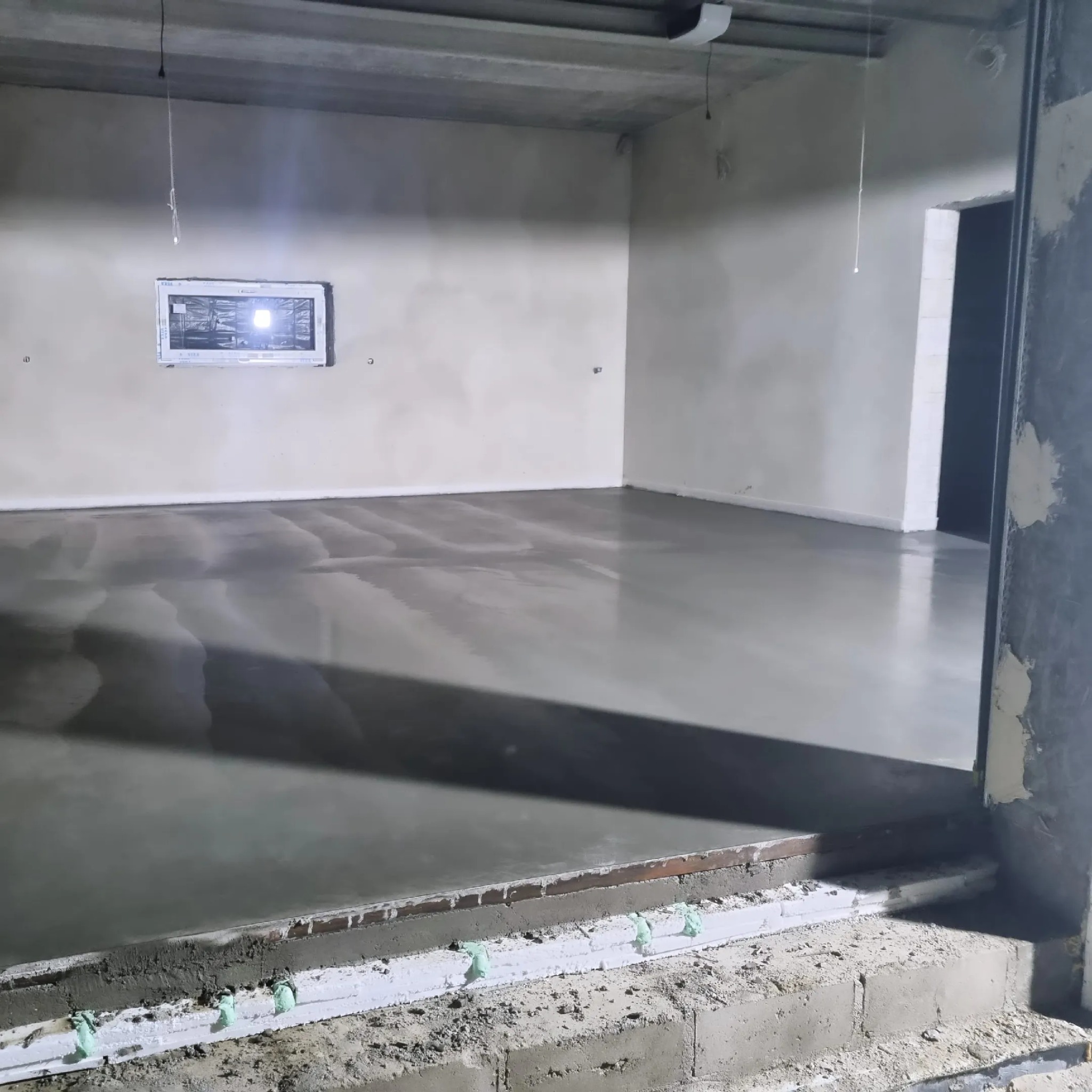 Świeżo wylana, błyszcząca posadzka jastrychowa w surowym wnętrzu z oknem i widoczną dylatacją przy ścianie, blokująca wylewkę.