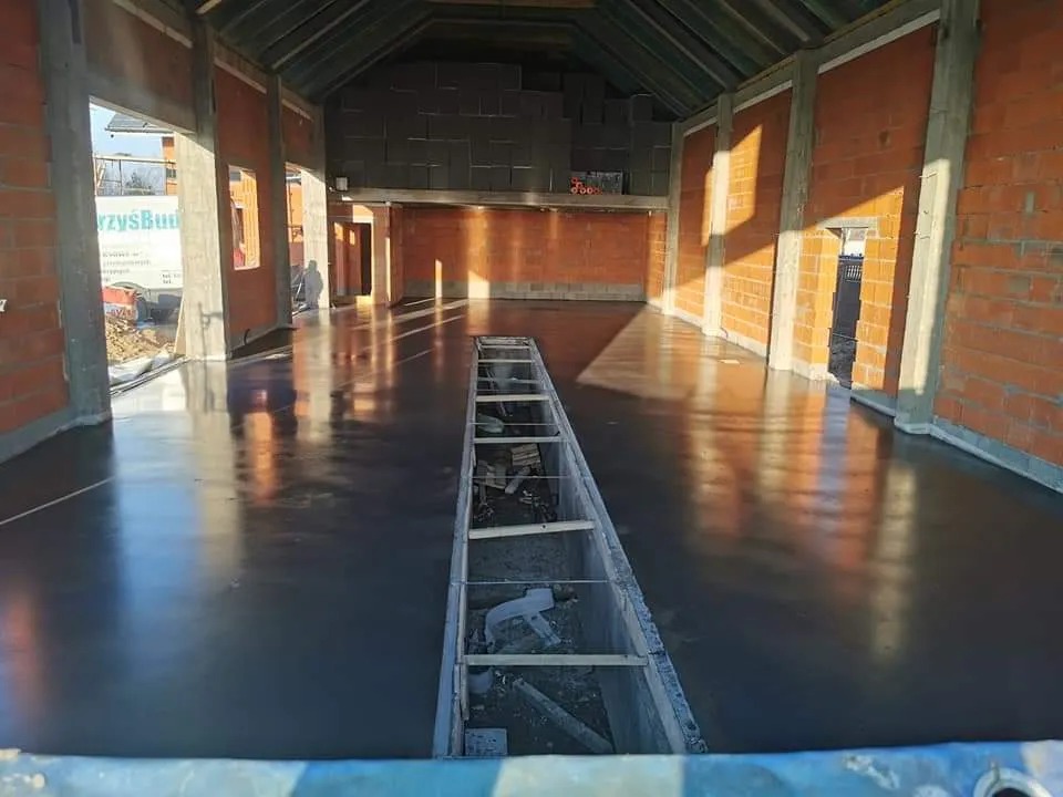 Świeżo wylana, gładka, ciemnoszara posadzka w surowym wnętrzu budynku w trakcie budowy, widoczne ceglane ściany i odkryta konstrukcja dachu, z kanałem instalacyjnym w podłodze.