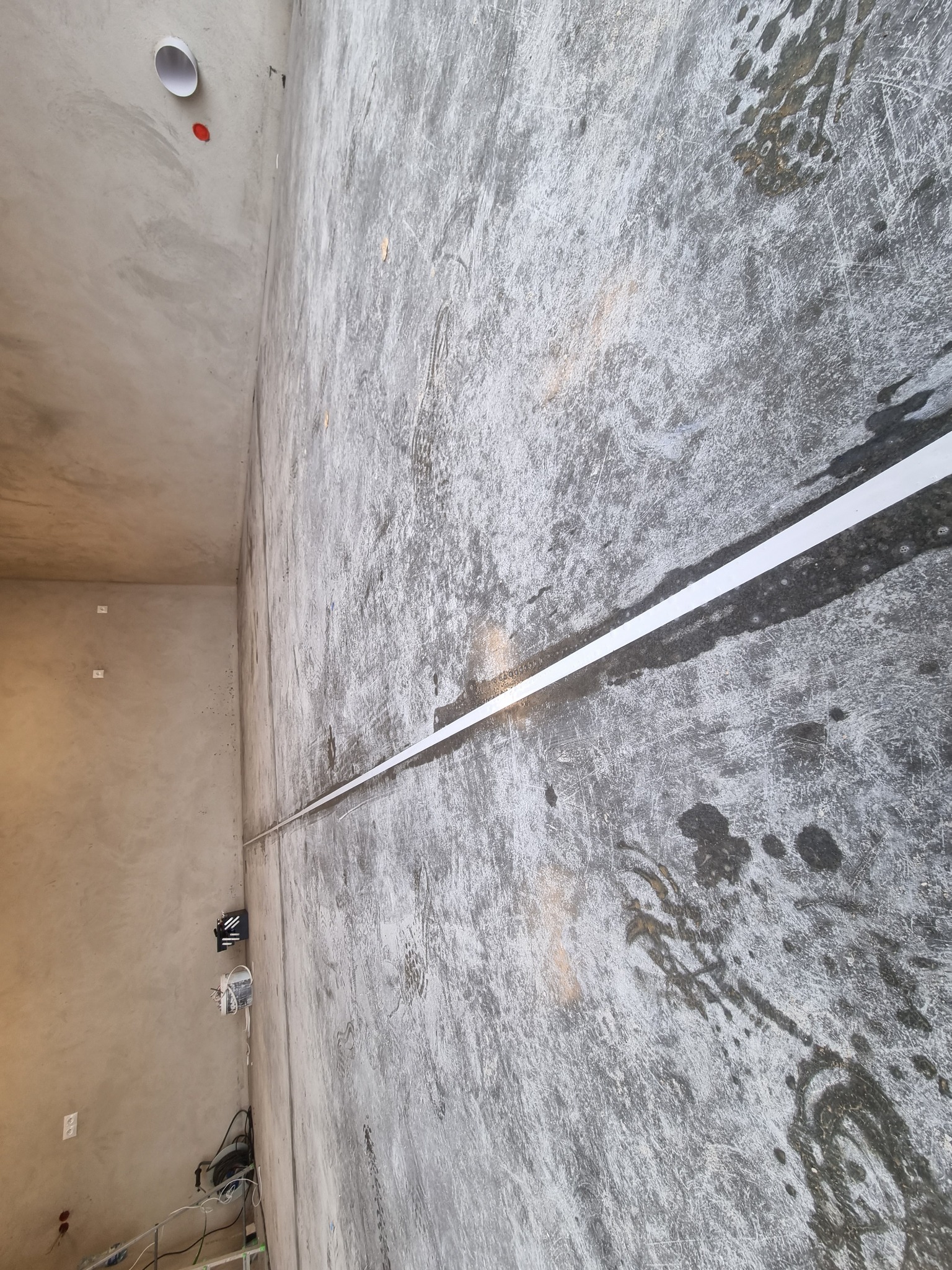 Surowa, szara posadzka w trakcie wylewania z widocznymi śladami po narzędziach i białym pasem oznaczającym linię, z elementami instalacji elektrycznej na ścianie w tle.