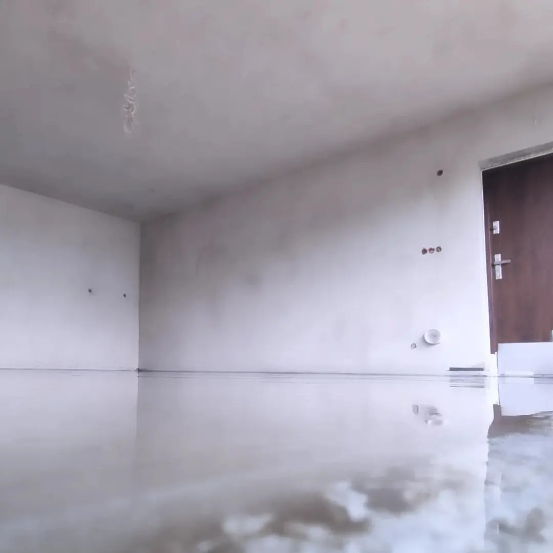 Świeżo wylana, gładka posadzka w pustym pomieszczeniu z białymi ścianami i brązowymi drzwiami. Powierzchnia podłogi odbija światło, tworząc efekt lustra.