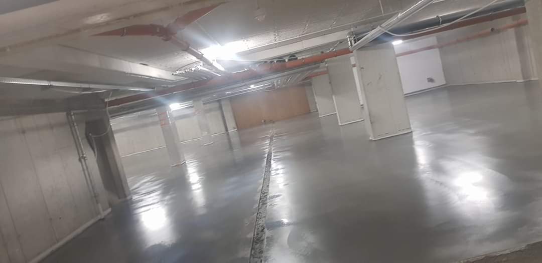 Gładka, szara posadzka przemysłowa w hali magazynowej z widocznymi słupami konstrukcyjnymi i instalacjami pod sufitem, z dylatacją pośrodku.