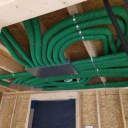 Zielone, karbowane rury wentylacyjne zamontowane pod sufitem budynku w stanie surowym, przymocowane metalowymi obejmami do drewnianych belek stropowych, widoczny fragment czarnej rury i otwór...