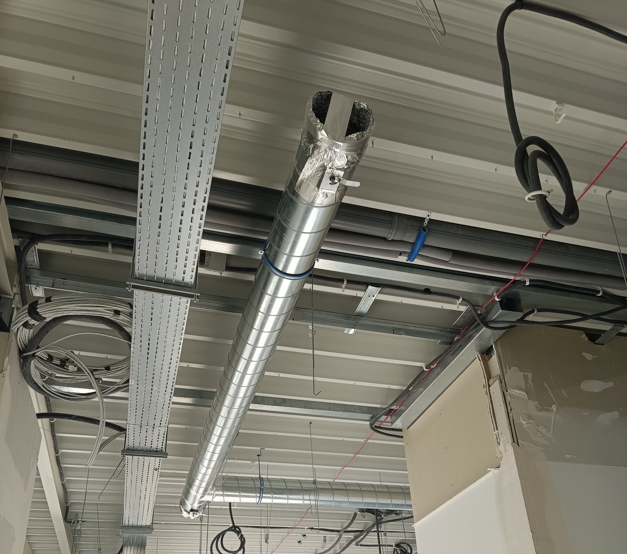 Widok z dołu na spiralny, metalowy przewód wentylacyjny pod sufitem, częściowo owinięty folią aluminiową, z widocznymi kablami i metalowymi elementami konstrukcyjnymi.