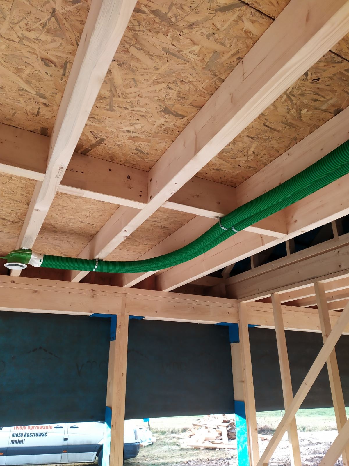Fragment instalacji wentylacyjnej z dwoma zielonymi, karbowanymi przewodami poprowadzonymi pod drewnianym stropem z płyt OSB w surowym stanie budynku.