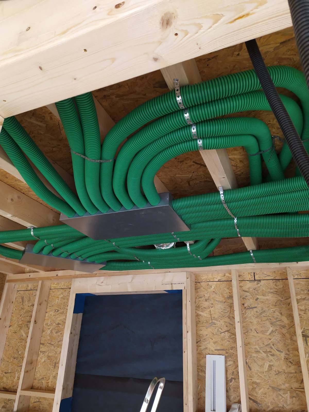 Zielone, karbowane rury wentylacyjne zamontowane pod sufitem budynku w stanie surowym, przymocowane metalowymi obejmami do drewnianych belek stropowych, widoczny fragment czarnej rury i otwór...