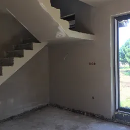 Wnętrze w trakcie prac wykończeniowych: ściany pokryte szarą gładzią, surowe schody betonowe, okno z widokiem na zieleń na zewnątrz, widoczne puszki elektryczne w ścianie.