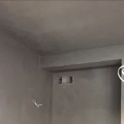 Świeżo nałożony tynk cementowo-wapienny na ścianie i suficie, widoczne dwa otwory w ścianie, fragment okna z profilem Salamander oraz zwisający kabel.