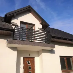 Czarna, pozioma balustrada na balkonie domu jednorodzinnego z brązową elewacją i ciemnym dachem, widziana z dołu w słoneczny dzień.