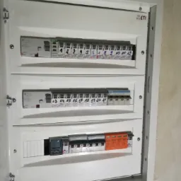 Otwarta skrzynka bezpiecznikowa z trzema rzędami wyłączników, oznaczonych numerami i symbolami, zamontowana na ścianie o nierównej fakturze.