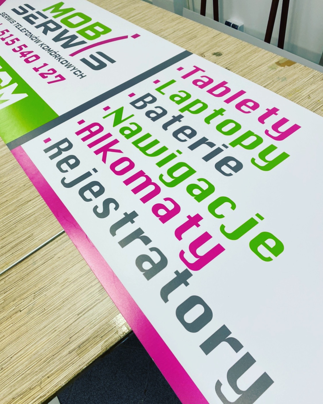 Wydrukowany baner reklamowy serwisu telefonów komórkowych z listą oferowanych usług, takich jak tablety, laptopy, nawigacje, na drewnianym blacie.