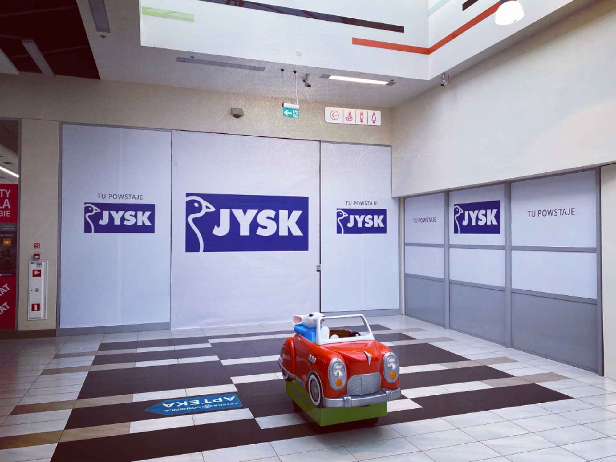 Okolicznościowa grafika z logo JYSK na białych panelach w przestrzeni handlowej, z zabawką - czerwonym autkiem - na pierwszym planie.